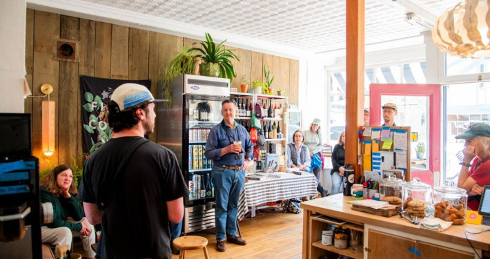 A group of people gather around a speaker giving a talk in the sunny 44 North cafe during Wings Waves and Woods 2025.